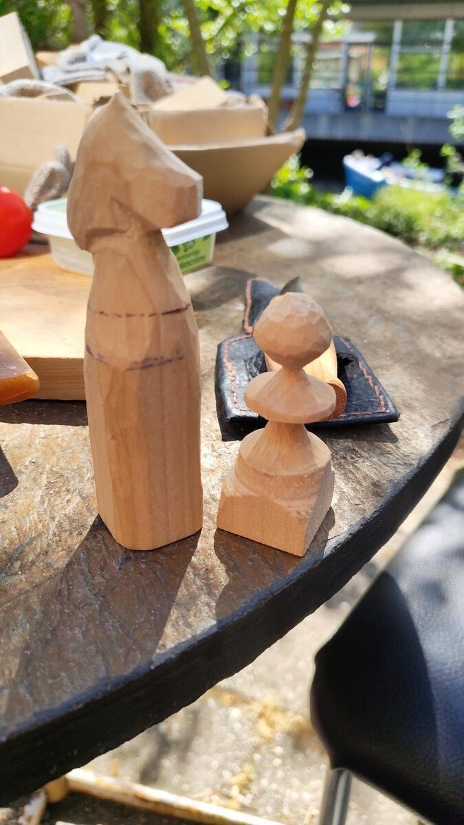 A roughed-out wooden knight, still showing pencil guidelines and tool facets, standing beside a finished pawn on a slate garden table in the sun. A leather tool roll and offcuts sit behind them.