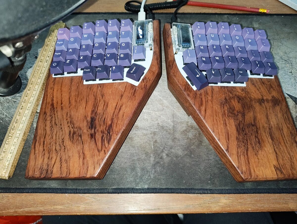 The two halves of a split ergonomic keyboard with purple keycaps, each mounted on a hand-cut wooden base, laid out on a workbench next to a folding ruler.